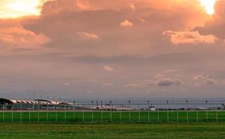 landskap av grönt gräsfält och staket av flygplatsen och vacker solnedgångshimmel. kommersiella flygplan parkerat vid förklädet av flygplatsen. Coronaviruset påverkade flygverksamheten. staket för säkerhet och säkerhet. foto