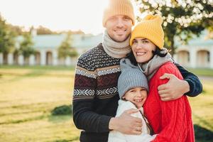 horisontellt porträtt av ung stilig man i gul hatt och varm stickad tröja, omfamna sin fru och dotter, posera vid kameran medan du står i parken. tre familjemedlemmar tillbringar helger tillsammans foto