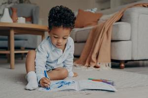 liten afrikansk pojke sitter på golvet och ritar med blå filtpenna foto