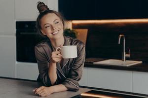 glad leende ung kvinna i brun satängpyjamas njuter av aromatiskt morgonkaffe i köket foto