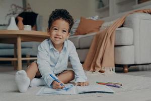 liten afro kid pojke sitter på golvet och ritar med färgglada filtpennor foto