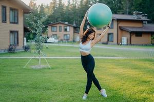 smal ung europeisk kvinna håller fitball över huvudet, klädd i beskuren topp och leggings, har gymnastikövningar utomhus, poserar på grön gräsmatta nära privat hus. människor, sport, träningskoncept foto