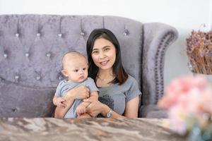 lycklig familj, söt asiatisk nyfödd baby med ung mamma leende lyckligt ansikte sittande soffa hemma. oskyldigt litet bedårande barn. mamma bär, tar hand med kärlek. föräldraskap, mors dag koncept. foto