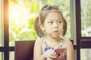 liten söt flicka tittar upp tänkande och håller smartphone, happy kid koncept foto