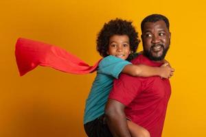 afrikansk far och son spelar superhjälte på dagtid. människor som har roligt gul bakgrund. begreppet vänlig familj. foto