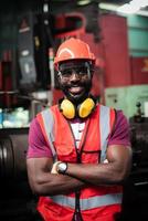 porträtt av glad afrikansk-amerikansk man arbetare har ovårdat skägg i säkerhetsuniform bär hjälm, glasögon, väst och handske i industriell tillverkare fabrik. foto