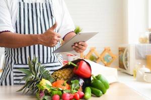 närbild av kockens hand använder surfplatta för att köpa frukt och grönsaker online. och det finns frukt och grönsaker och papperspåsar på köksbordet.shopping online och leveranskoncept. foto