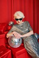 senior gråhårig kvinna i solglasögon och silver klänning med disco boll på festen på röda gardiner bakgrund. fest, firande, senior ålder koncept foto