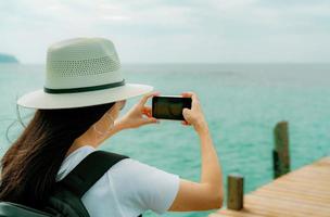 ung asiatisk backpacker kvinna bär hatt använda smartphone ta foto på piren. sommarsemester på tropisk paradisstrand. glad hipster flicka resa på semester. kvinna njuta och koppla av livet. sommarvibbar.