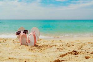 rosa och vita sandaler, solglasögon på sandstranden vid havet. casual mode stil flipflop och glasögon vid havet. sommarsemester på den tropiska stranden. rolig semesterresa på sandstranden. sommartid. foto