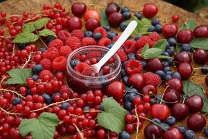 vit sked med bärsylt i glaset. blåbär, hallon, körsbär och vinbär på träbordet i trädgården. foto