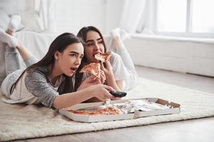 fritid. systrar äter pizza när de tittar på tv medan de ligger på golvet i det vackra sovrummet på dagtid foto
