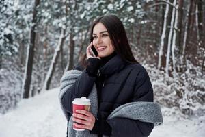 ringer till kompisen. flicka i varma kläder med en kopp kaffe har en promenad i vinterskogen foto
