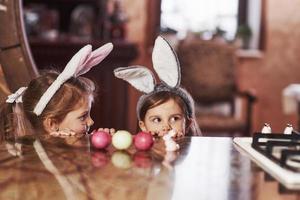 söta barn. roliga två systrar i kaninöron gömmer sig nära bordet med målade ägg foto