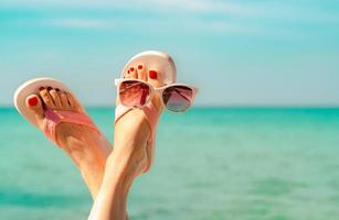 upp och ner kvinna fötter och röd pedikyr bär rosa sandaler, solglasögon vid havet. rolig och glad mode ung kvinna koppla av på semester. chill out tjej på stranden. kreativt för resebyrån. helgresor. foto