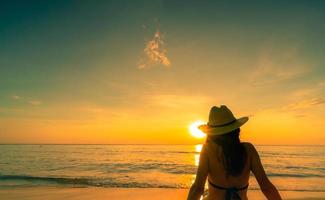 bakifrån av asiatisk kvinna bär bikini och halmhatt sitta på sandstranden, slappna av och njut av semestern på den tropiska paradisstranden. kvinna titta på vacker solnedgång. sommarsemester. resa ensam. sommarvibbar. foto