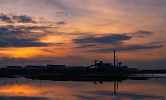 landskap av fabriksindustribyggnader med mörkblå och orange solnedgångshimmel reflektion på vatten i floden. lagerbyggnad på natten. ren miljö runt fabriken. fabriken stängd. foto