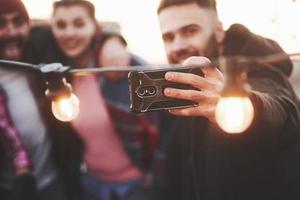fokuserad bild. grupp unga glada vänner har roligt, kramar varandra och tar selfie på taket med dekorerade glödlampor foto