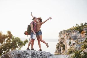 två turist man och kvinna med ryggsäckar står på toppen av klippan och njuter av soluppgången. resande berg och kust, frihet och aktivt livsstilskoncept foto