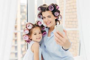 attraktiv liten unge med positivt uttryck, charmigt leende står nära sin mamma, gör selfie med modern mobiltelefon, posera mot fönsterbakgrund i rummet. föräldraskap och skönhetskoncept foto