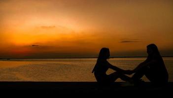 silhuett av två söta flickor sitter och håller hand och har kul tillsammans vid havet i solnedgången med orange himmel. sommarsemester och ryggsäcksresor koncept. foto