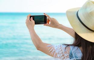 asiatisk hipster kvinna bär hatt använder smartphone ta foto av vackra havet. sommarsemester på tropisk paradisstrand. glad hipster flicka resa på semester. kvinna njuta och koppla av. sommarvibbar.