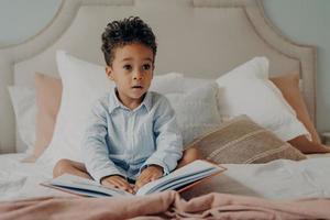 söt lockigt hår afroamerikansk liten pojke sitter på sängen med bok foto