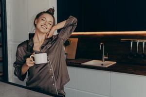 vacker avslappnad ung kvinnlig modell i mysig pyjamas med en kopp kaffe i köket foto