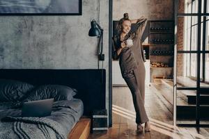 ung ganska smal kvinna som bär brun satin pyjamas håller kaffekoppen medan du står i sovrummet foto