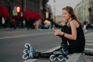 utomhusbild av sportig aktiv ung kvinna klädd i aktivt slitage bär rollerblades kontrollerar meddelandeinnehåll på smartphone poserar mot suddig stadsbakgrund på asfalt leder hälsosam livsstil foto