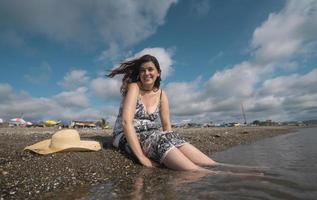 vacker glad ung latinamerikansk kvinna sitter på kanten av stranden bredvid sin hatt, klädd i en svartvit klänning under en solig morgon foto