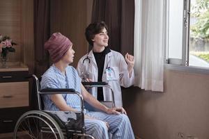 uniformerad ung kaukasisk kvinnlig terapiläkare som uppmuntrar manlig patient i rullstol vid fönstret för att stödja och motivera tillfrisknandet, cancersjukdom efter kemomedicinsk behandling på sjukhusets slutenrum. foto