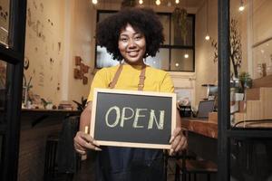 en afrikansk amerikansk kvinnlig startbarista står vid casual cafédörren, tittar på kameran och visar öppen skylt, glada och glada leenden med jobb i kaféservice och nya företagare. foto