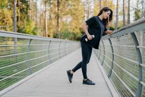 idrottskvinna håller hantlar och tränar utomhus, klädd i aktivt slitage, poserar på bron och tittar ner, skog i bakgrunden. sportig frisk stark kvinna tränar muskler under träning foto