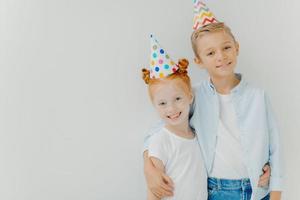 glad liten ingefära fräknig tjej och liten pojke omfamnar och ler glatt, bär festhattar, njut av trevlig tid tillsammans, isolerade över vit bakgrund, kopiera utrymme. barn och festligt evenemang koncept foto