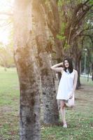 vacker asiatisk kvinna i vit klänning koppla av och ler i naturparken. Thai flicka eller kinesisk flicka njuta på semester med solljus i trädgården foto