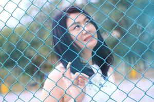 vacker asiatisk kvinna i vit klänning som sitter och ler i naturparken. Thai flicka eller kinesisk flicka njuta på semester med solljus i trädgården. stängt fokus på blå galler stål staket. foto