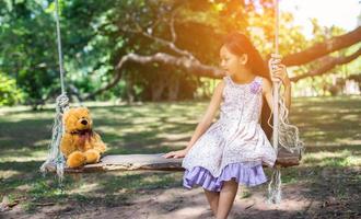 söt liten flicka sitter gungor, nallebjörn sitter med henne, liten flicka pekade på trädet. foto