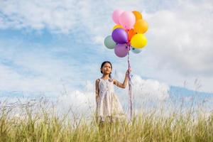 söt liten flicka håller färgglada ballonger på ängen mot blå himmel och moln, sprider händer. foto