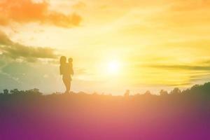 en siluett av ett lyckligt ungt flickbarn armarna på sin kärleksfulla mor för en kram, framför solnedgången på himlen en sommardag. foto