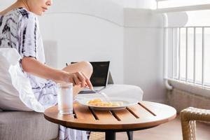 frisk kvinna sitter på en soffa och äter färsk frukt medan du arbetar på laptop. foto