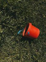röd leksakshink med trasigt blått handtag liggande på gräs, vertikalt foto, varma färger. plastleksak för barn för att leka med sand eller vatten, trädgårdsredskap foto