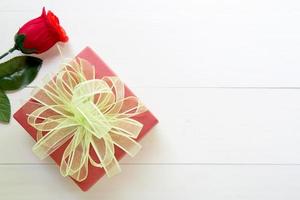 Presentgåva med röd rosblomma och presentförpackning med rosettband på träbord, 14 februari kärleksdag med romantiska, alla hjärtans semesterkoncept, ovanifrån. foto