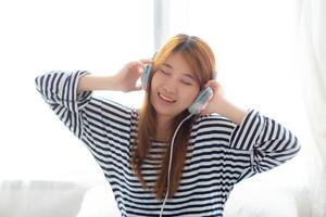 vacker asiatisk ung kvinna njuta och roligt lyssna musik med hörlurar sitter i sovrummet, flicka koppla av med hörlurar, fritid och teknik koncept. foto