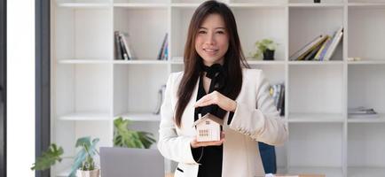 miniatyr hus i händerna på en asiatisk kvinna fastighetsmäklare bostadslån som arbetar på kontoret. tittar på kameran. foto