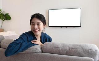 framför en glad asiatisk kvinna som tittar på kameran. bakom den vita tv:n i soffan i vardagsrummet hemma. foto