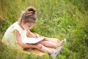 flicka i en gul klänning sitter i gräset på en filt på ett fält och läser en pappersbok. internationella barndagen. sommartid, barndom, utbildning och underhållning, stugkärna. kopieringsutrymme foto