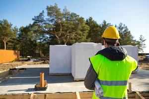 byggaren pratar i telefon. arkitekt i en skyddande gul hjälm och en signalväst står ryggar framför byggarbetsplatsen till ett hus med grund och lättbetongblock. foto