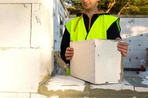Byggaren håller ett block av cellbetong i sina händer - murverket på husets väggar. byggnadsarbetare i skyddskläder - en hjälm och en reflexväst. foto