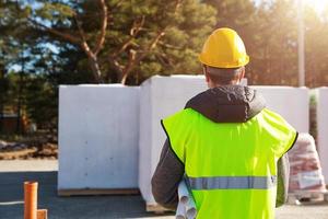 projektarkitektens rygg är på byggarbetsplatsen för ett hus med grunden lagd och blocken levererade. byggnadsarbetare i en skyddande gul hjälm och en signalväst. håna foto